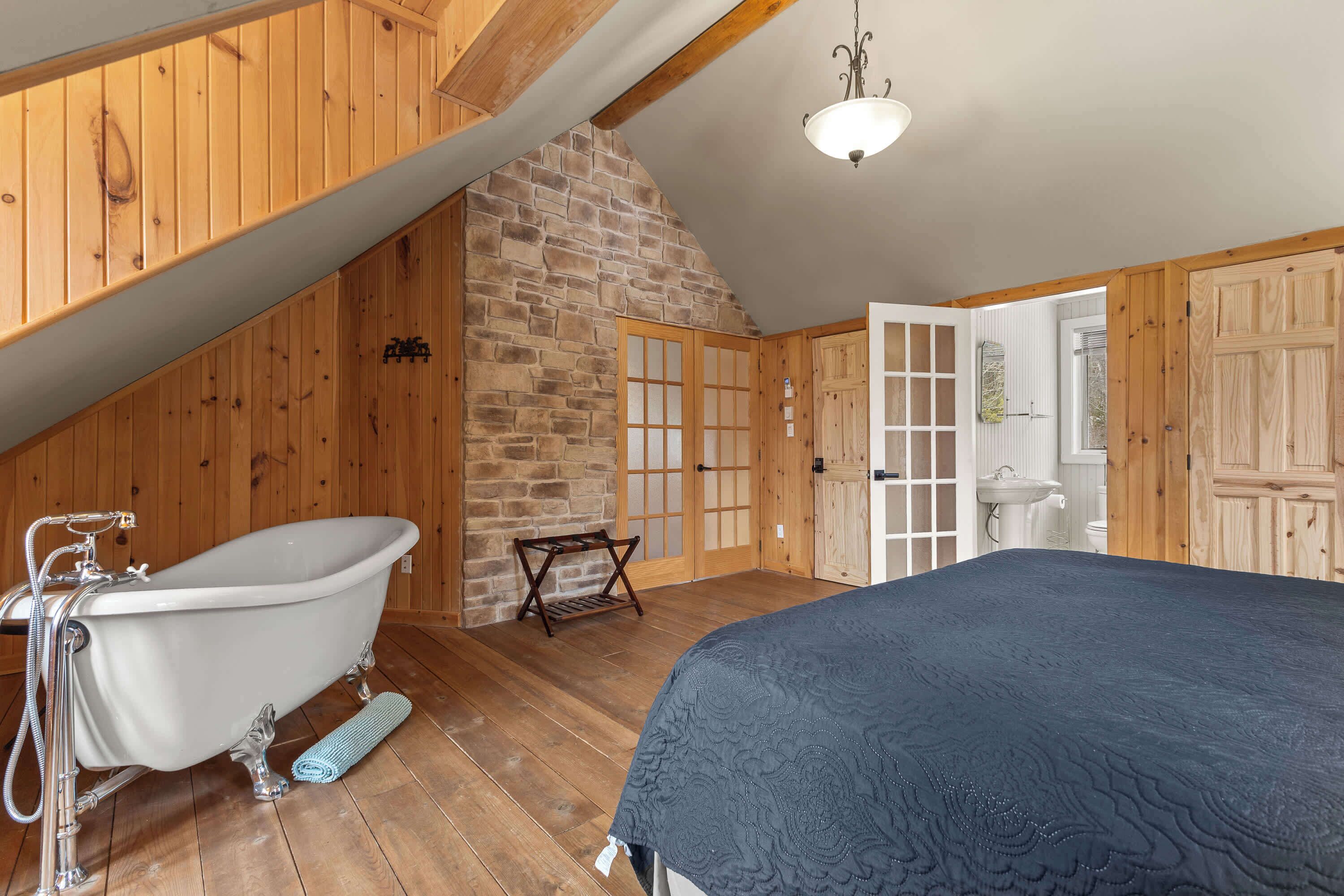 A room with a wooden floor, a white bathtub, a bed with a dark quilt, a stone wall, and wooden doors.