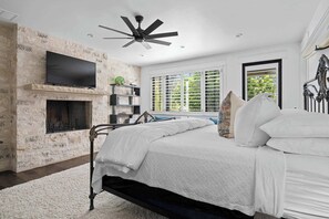 Une chambre avec un mur en pierre, une cheminée, un lit avec une literie blanche, un ventilateur de plafond et un téléviseur fixé au mur.