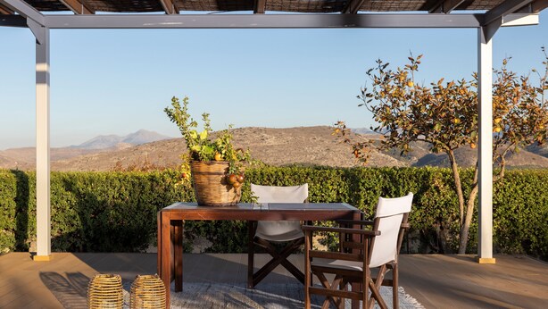 Eine Terrasse im Freien mit einem Holztisch und Stühlen, einem Korb voller Früchte und Blick auf Berge und einen Heckenstreifen.