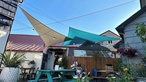 A patio area with a wooden table and chairs, a teal shade sail, and various potted plants.