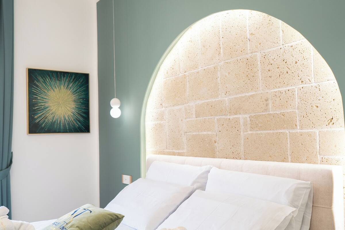 Uma cama bem arrumada com lençóis brancos e almofada verde, uma obra de arte na parede e uma cabeceira arqueada peculiar.