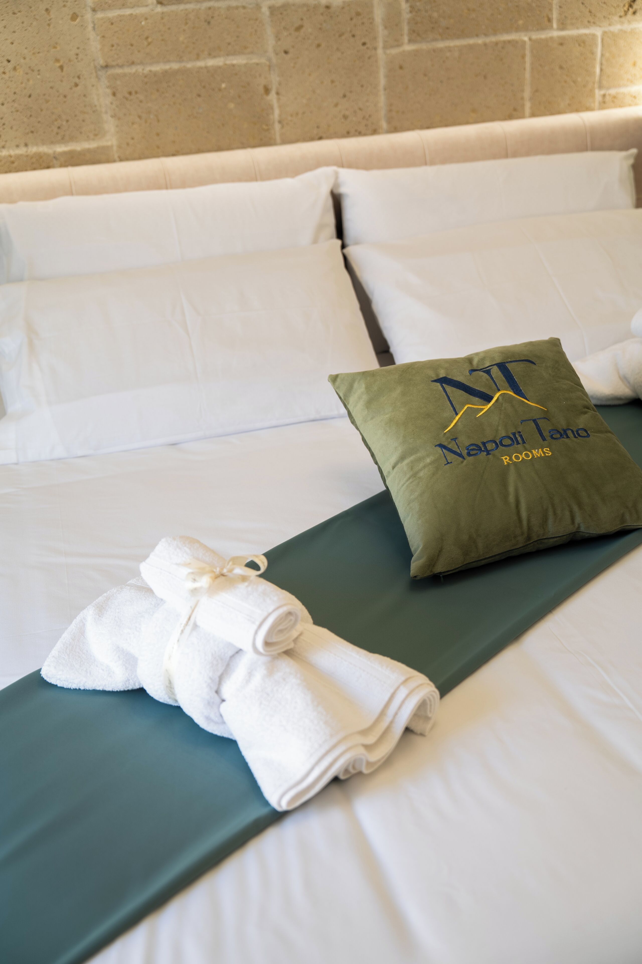 A neatly made bed with white linens, a green pillow with a logo, and a rolled white towel on a green bed runner.