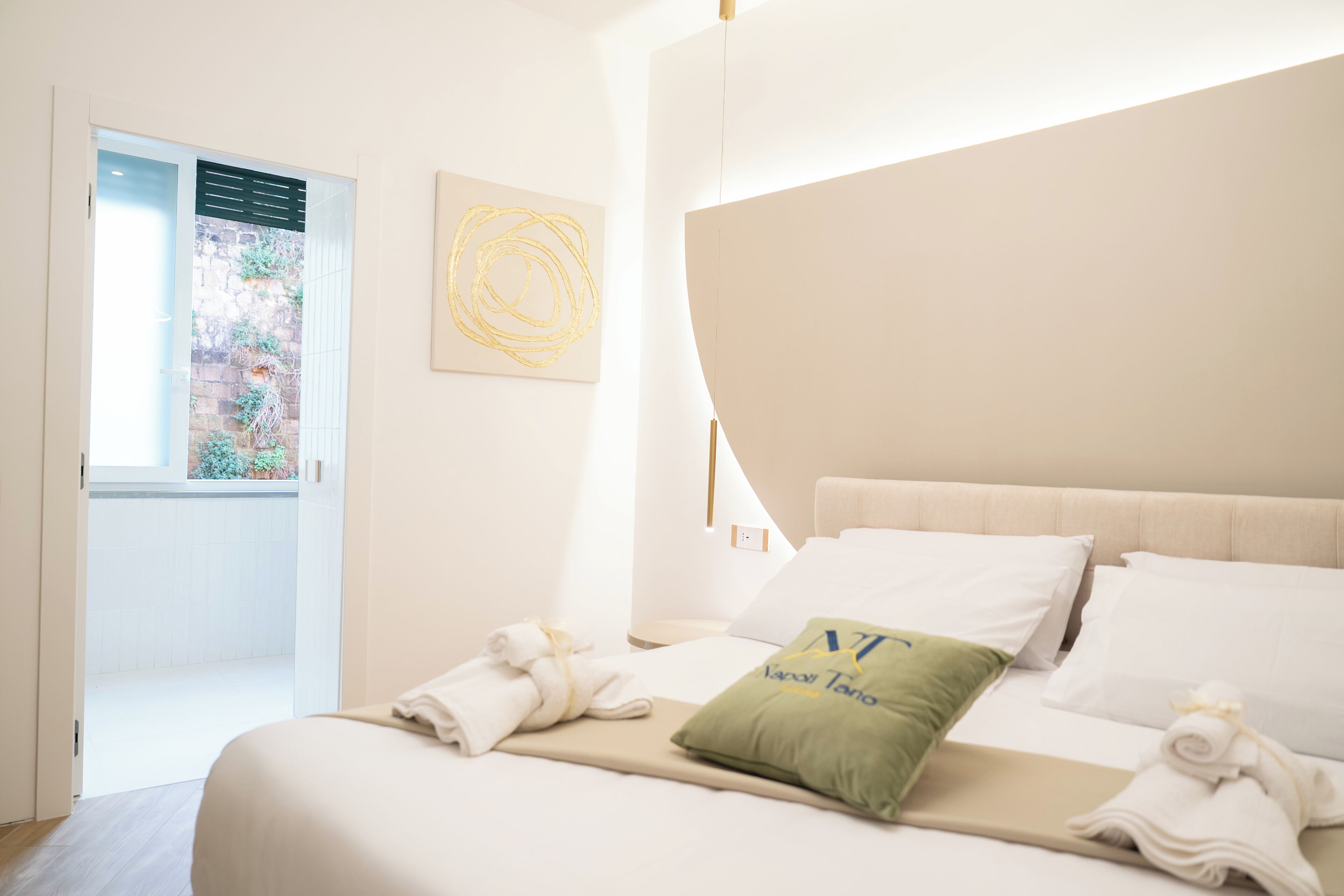 A modern hotel room with a bed, a green pillow, a towel, a wall art piece, and a window with a view of greenery.