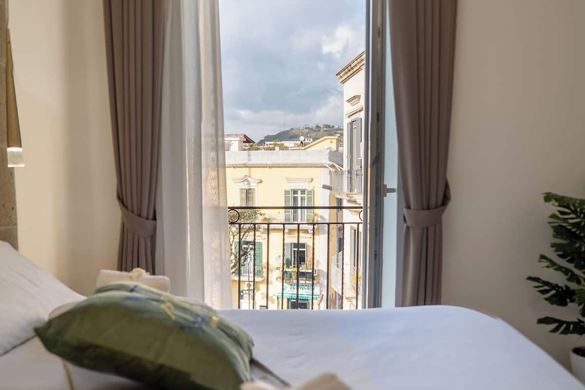 Quarto de hotel com cama, travesseiro verde, toalha enrolada e vista para prédios por uma janela aberta.