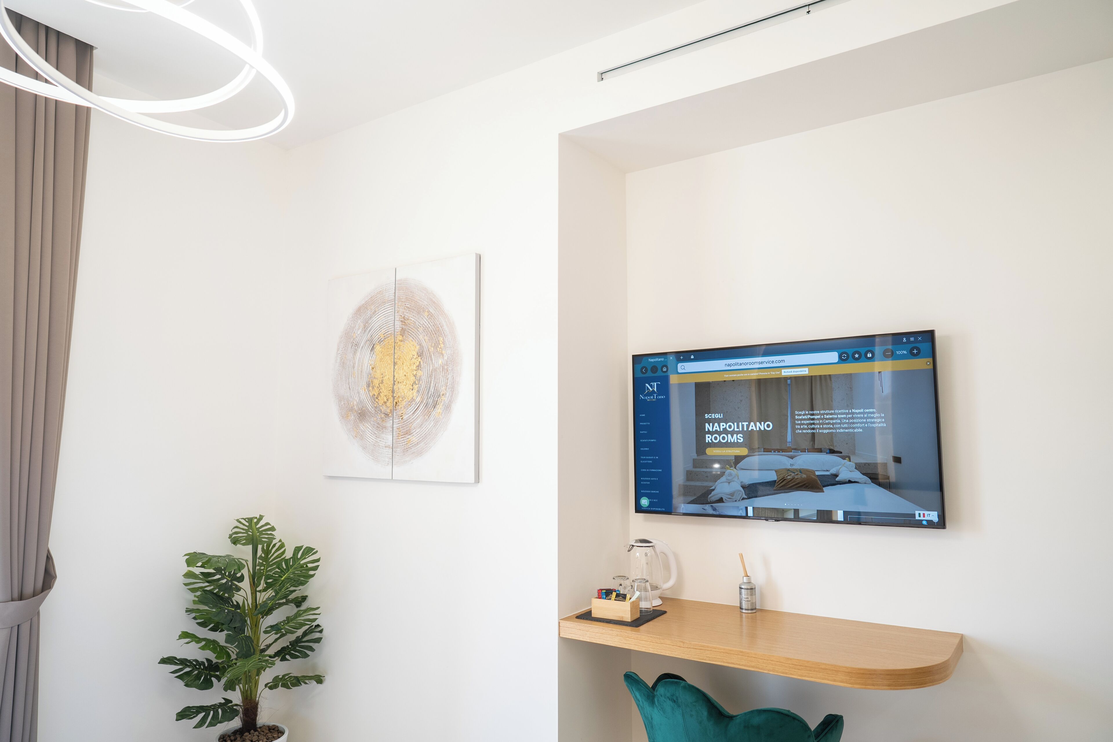 A modern hotel room with a large flat-screen TV displaying a website, a potted plant, a wooden shelf with a coffee maker, and a framed abstract artwork.