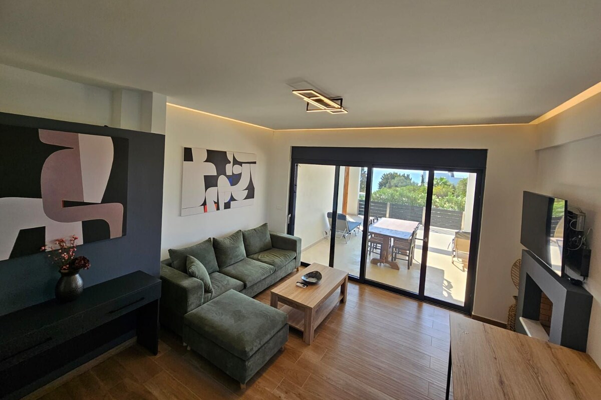A modern living room with a sofa, coffee table, and a TV. There is a sliding glass door leading to a patio area.