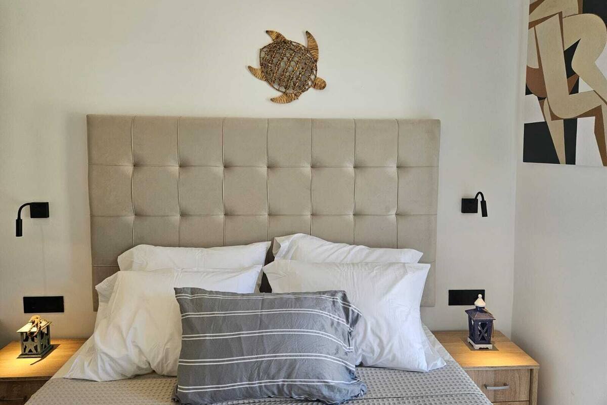 Un dormitorio con cabecera capitonada, cama con almohadas blancas y a rayas, decoración de tortuga montada en la pared y mesita de noche con un farol.