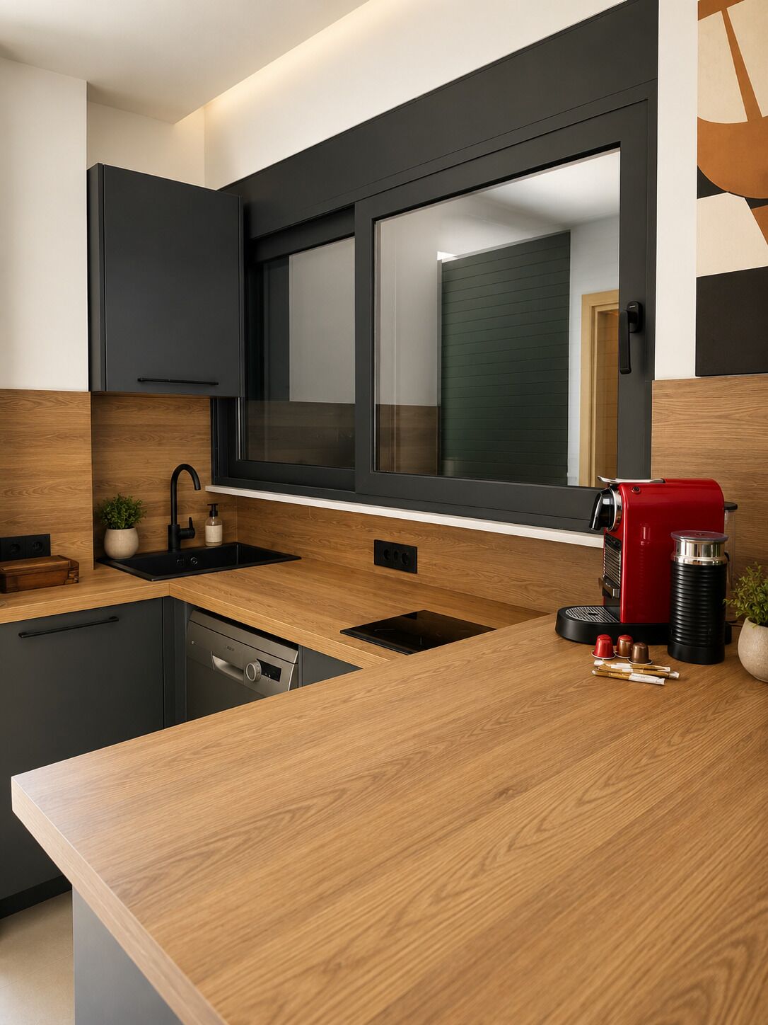 Una cocina moderna con encimera de madera, armarios negros, una cafetera roja y un ventanal sobre el fregadero.