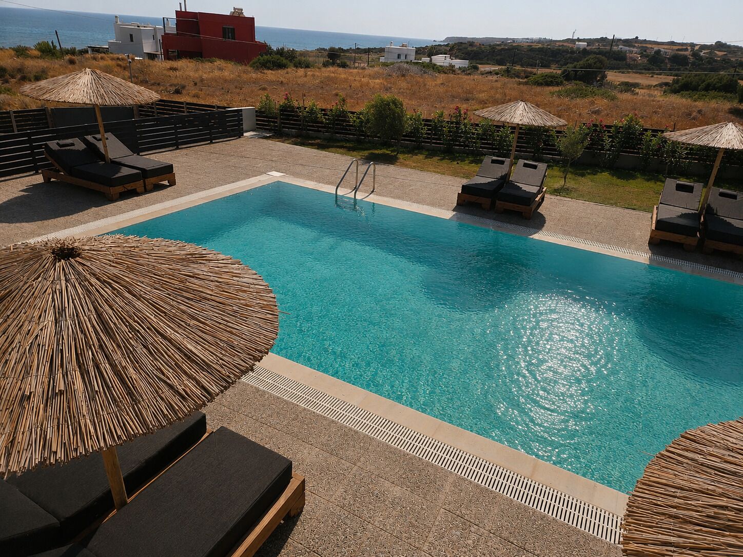 A pool area with lounge chairs and umbrellas, surrounded by a landscaped garden and a view of the sea.