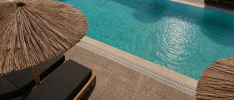 A pool area with lounge chairs and umbrellas, surrounded by a landscaped garden and a view of the sea.