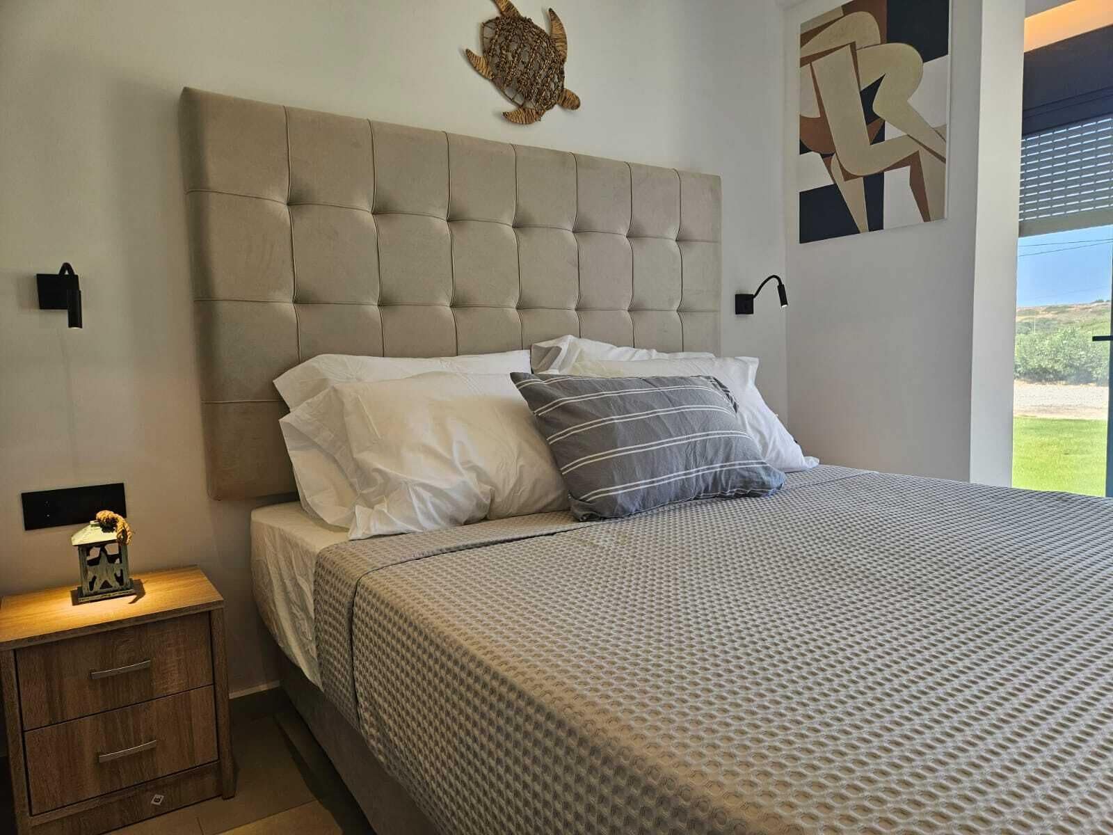 Une chambre à coucher avec une tête de lit capitonnée, un lit recouvert d’une literie à motifs, une table de chevet avec une lampe et une décoration murale représentant une tortue.
