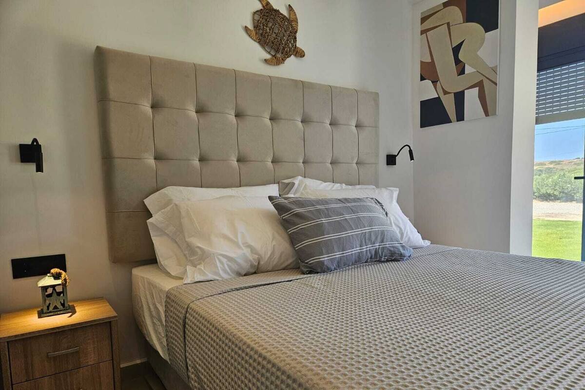 Un dormitorio con cabecera capitonada, cama con ropa de cama estampada, mesita de noche con lámpara y una decoración de tortuga colgada en la pared.