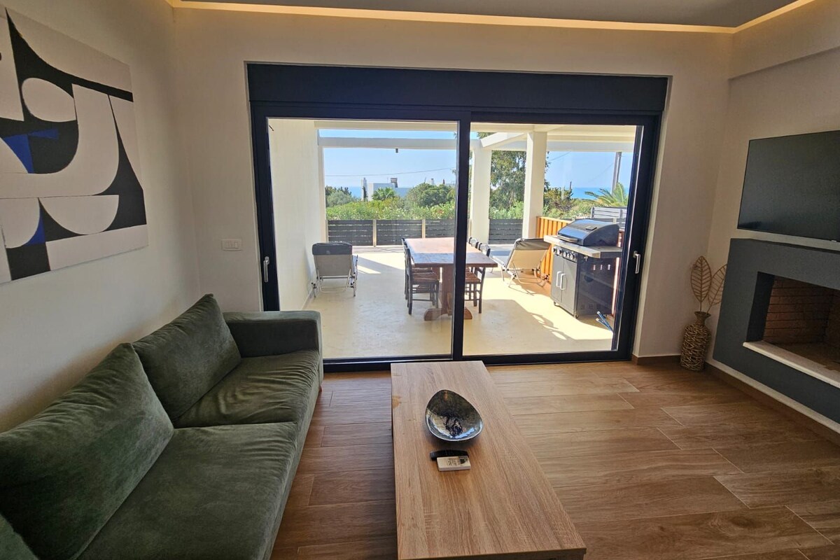 A modern living room with a sofa, a coffee table, and a sliding glass door leading to an outdoor patio with a grill and dining area.