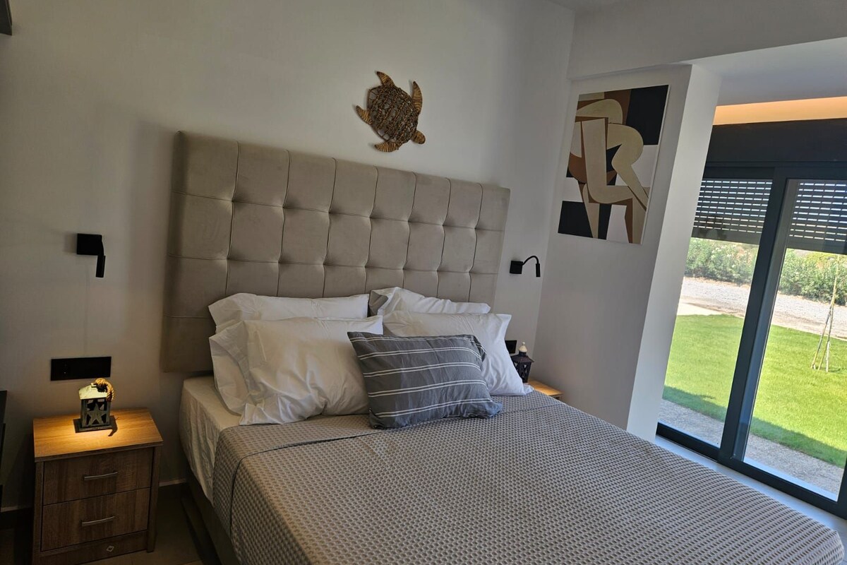 A bedroom with a tufted headboard, a bed with patterned bedding, a nightstand with a lamp, and a view of a green lawn through a large window.