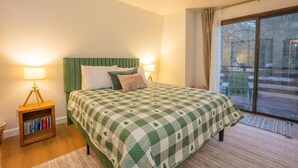 A bedroom with a checkered bedspread, a green headboard, bedside lamps, a nightstand with books, and a view through a large window. - Big Bear Moonridge Escape (Big Bear Lake)