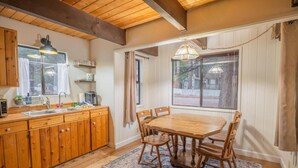 A wooden kitchen with a dining table and chairs, a window with curtains, and a hanging light fixture. - Big Bear Moonridge Escape (Big Bear Lake)