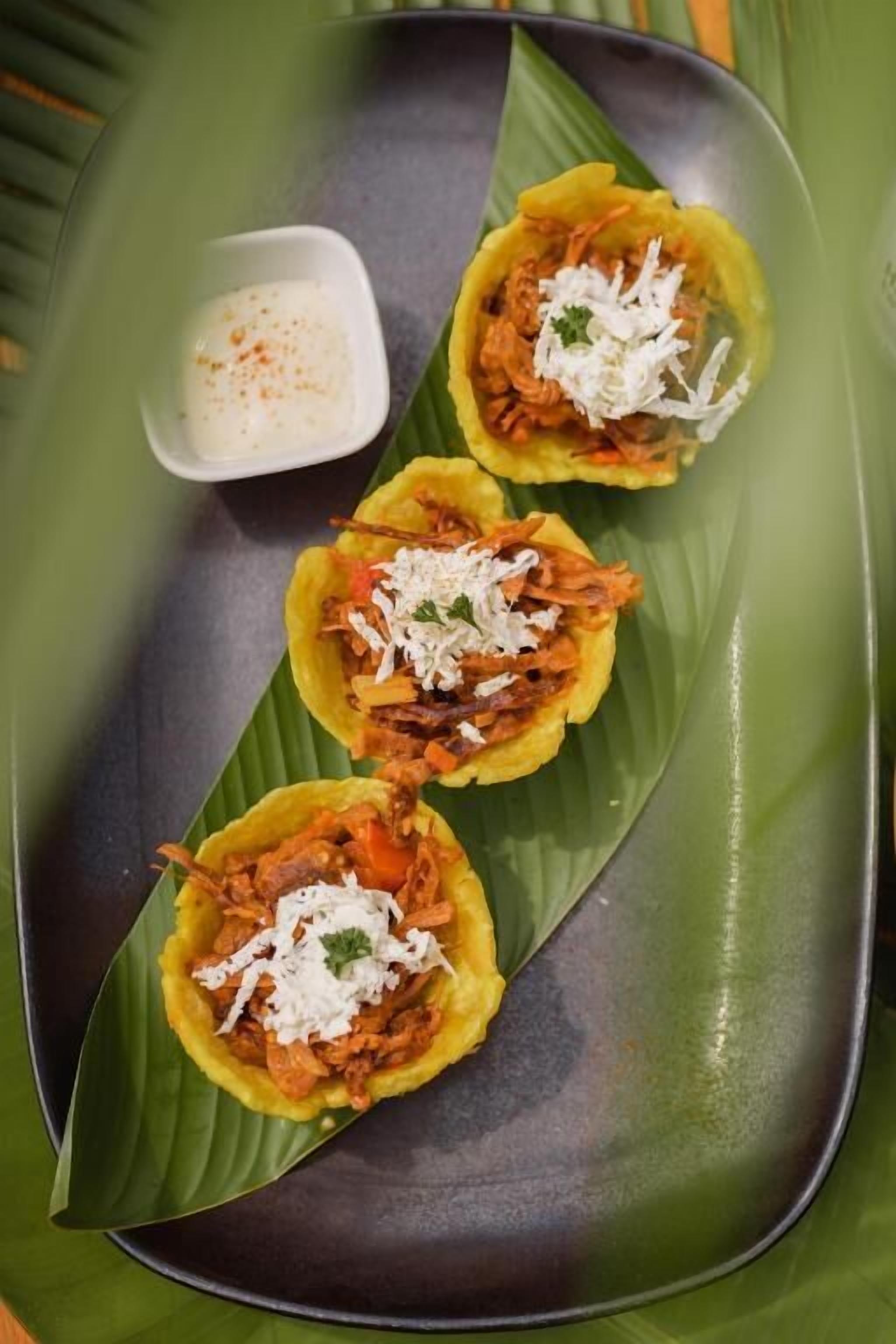 A plate with three tacos on a banana leaf, a small bowl of sauce, and a dark plate.