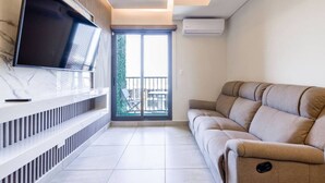 A modern living room with a flat-screen TV, a sofa, a balcony with a table and chairs, and an air conditioning unit.