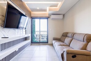 A modern living room with a flat-screen TV, a sofa, a balcony with a table and chairs, and an air conditioning unit.