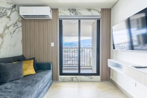 A modern living room with a sofa, a flat-screen TV, an air conditioning unit, and a sliding glass door leading to a balcony with a city view. - 1 Queen, 1 Sofa Bed, Wifi, Gym, Pool - 4 people (Monterrey)