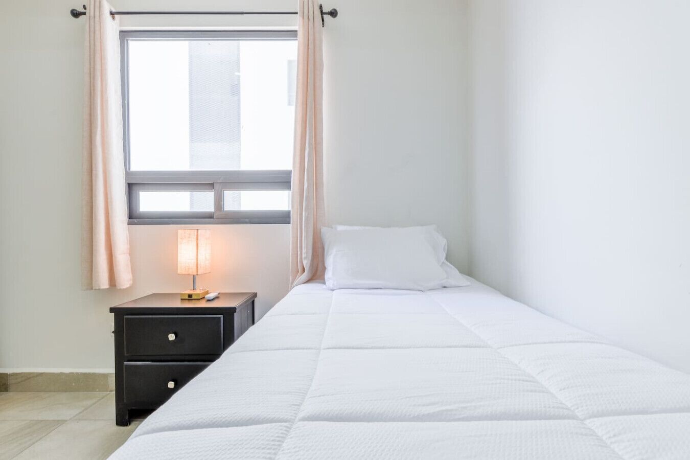 Ein Einzelbett mit weißer Decke, ein Nachttisch mit Lampe und ein Fenster mit Vorhängen.