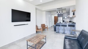 Un salon moderne doté d’un téléviseur à écran plat, d’une table basse en bois, d’un canapé en cuir noir et d’un coin cuisine avec des tabourets de bar.