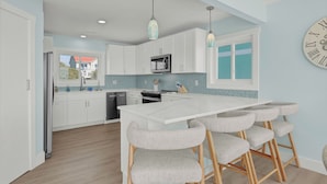 A modern kitchen with white cabinets, a central island with bar stools, a clock on the wall, and a view of the outside.