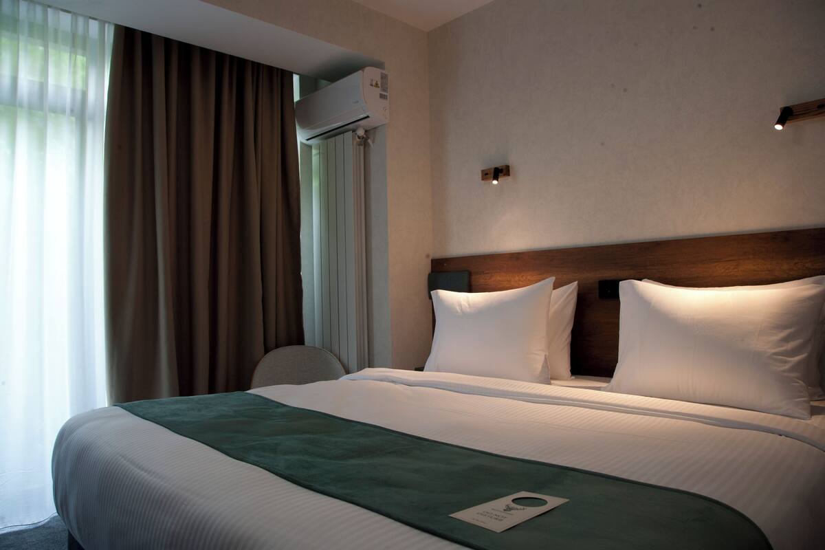 Una habitación de hotel con cama, una silla, ventana con cortinas, equipo de aire acondicionado y una lámpara fijada a la pared.
