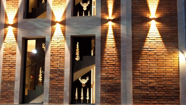Fachada de edifício com detalhes em tijolo, colunas verticais e janelas iluminadas.