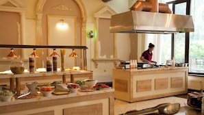 Buffet montado com diversos alimentos, um cozinheiro preparando refeições e vista para o exterior através de grandes janelas.