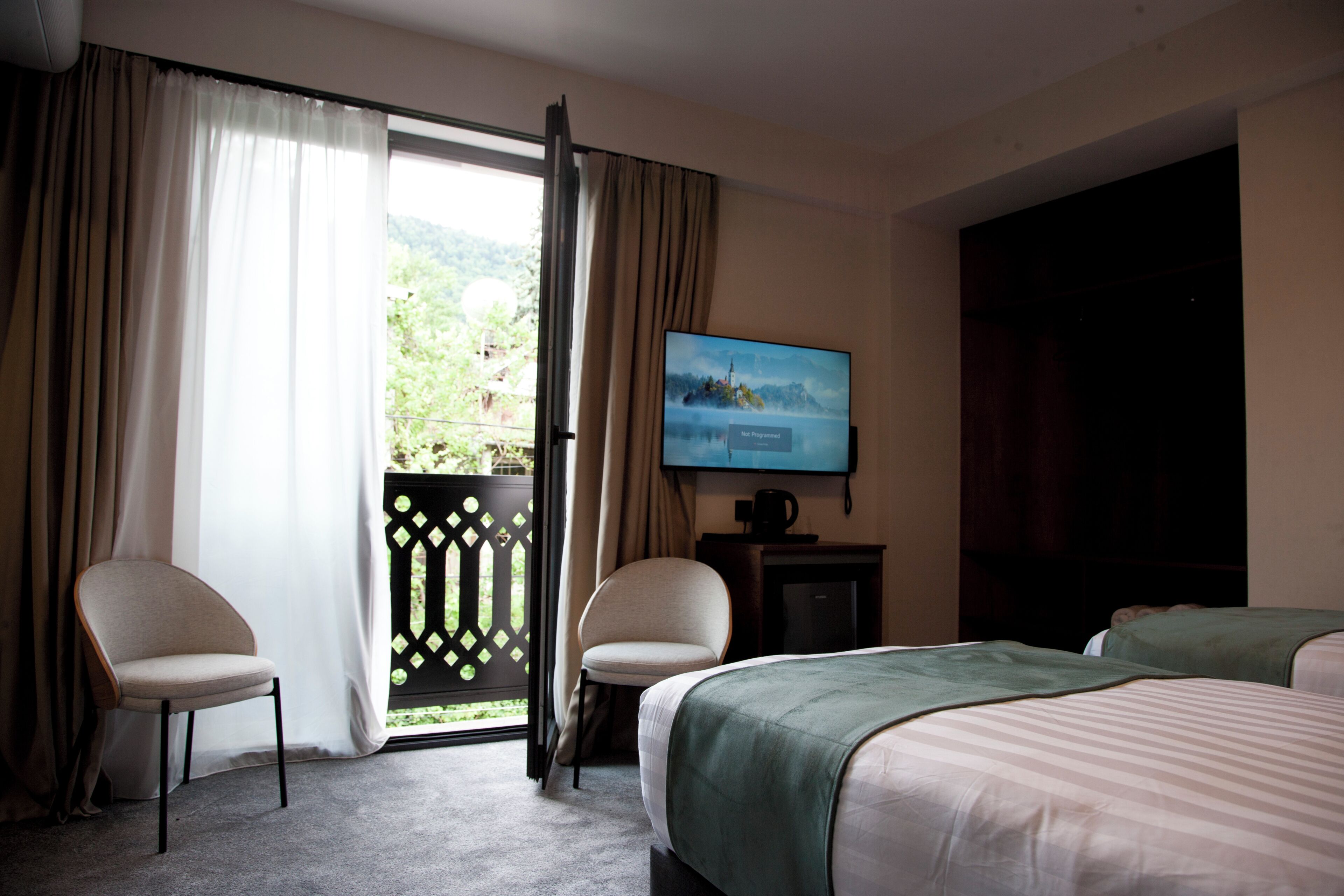A hotel room with a bed, two chairs, a TV, and a view of greenery through a window.