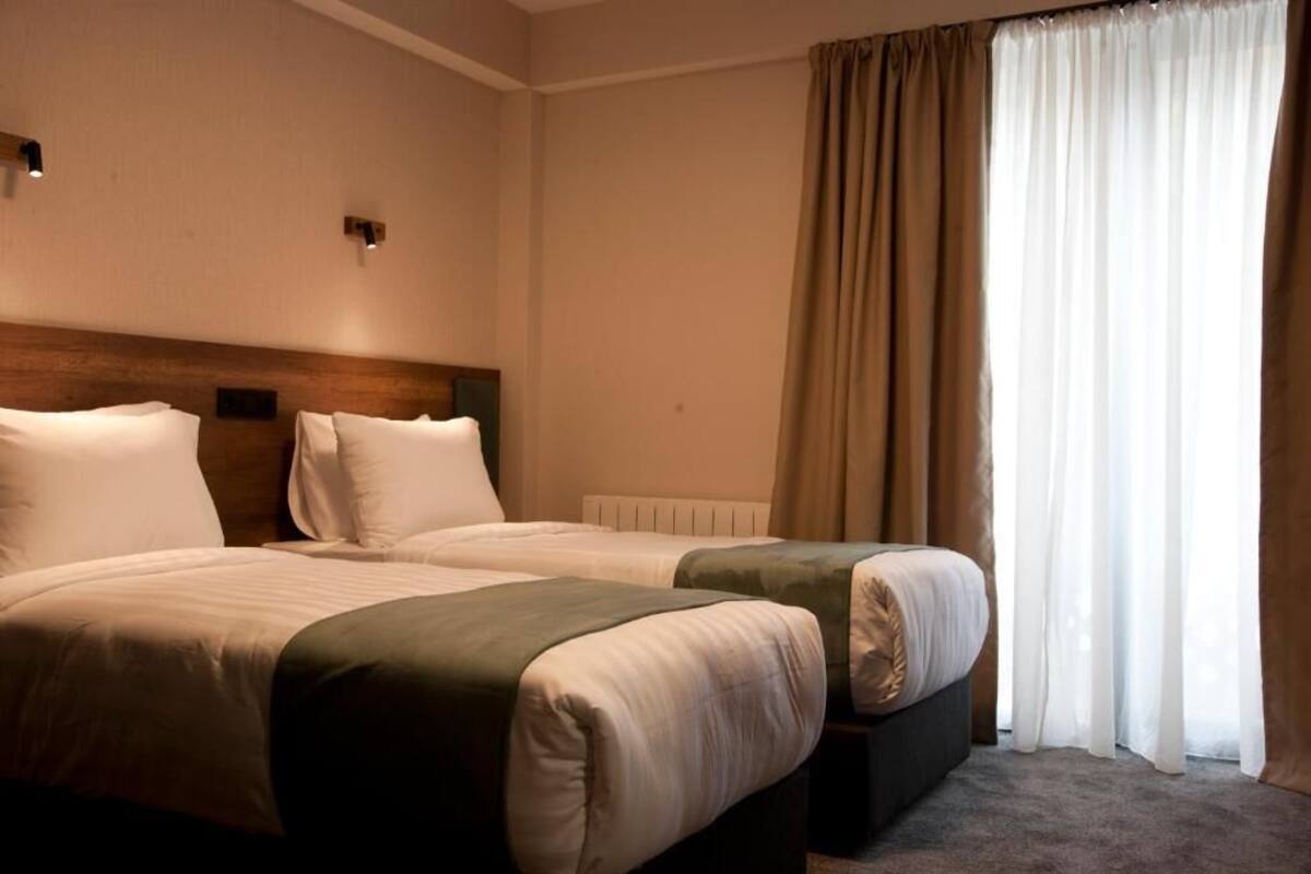 Una habitación de hotel con dos camas, un cabecero de madera, un radiador y un ventanal con cortinas.