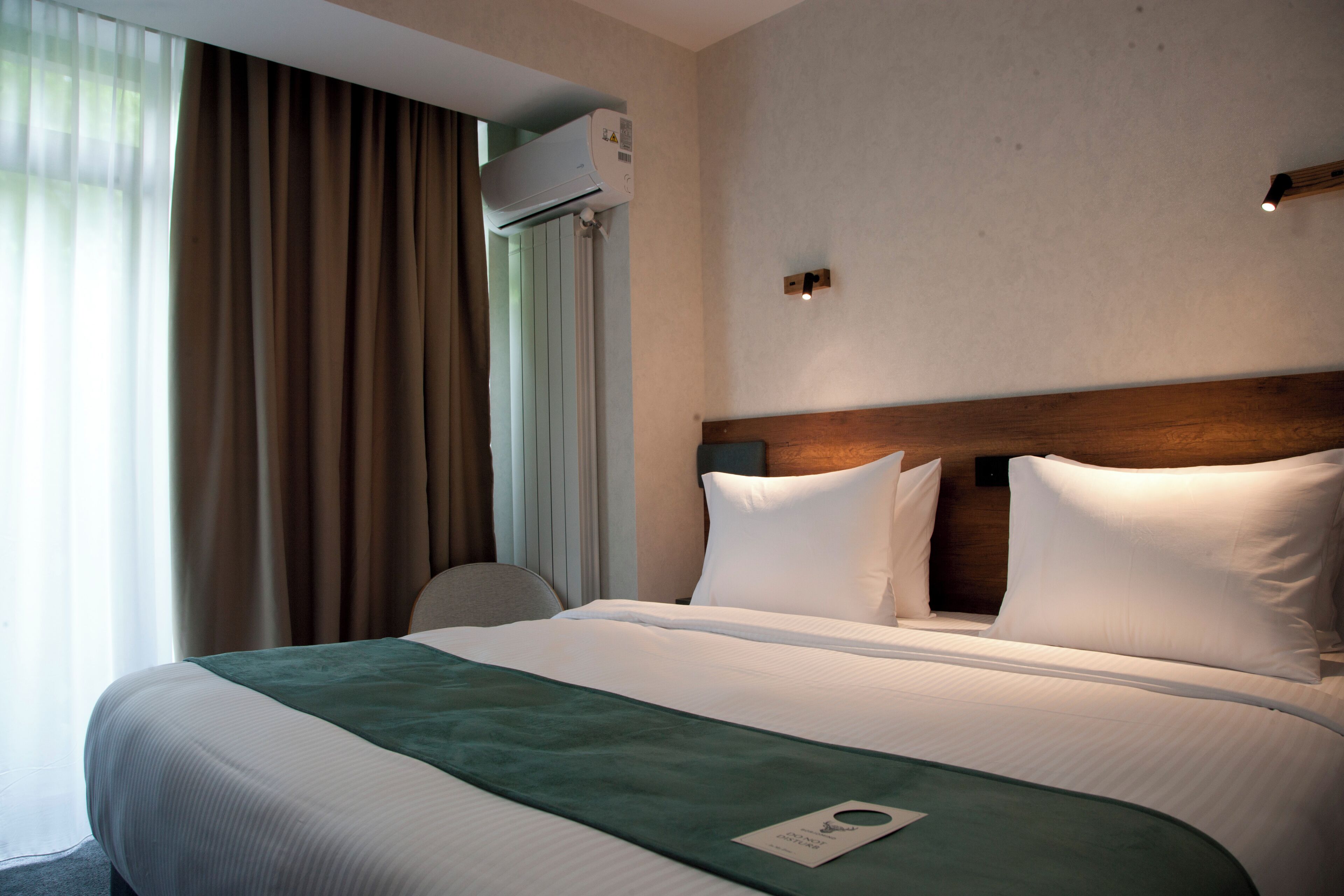 Una habitación de hotel con una cama grande, un cabecero, una ventana con cortinas, un equipo de aire acondicionado y una lámpara de pared.