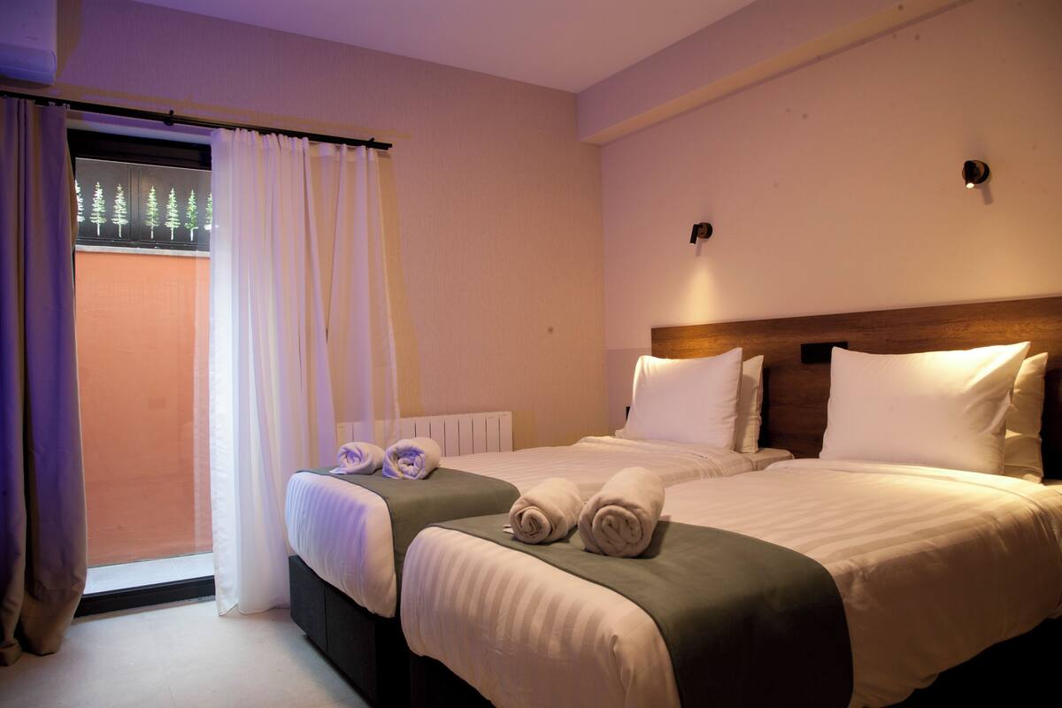 Habitación de hotel con dos camas, una ventana con cortinas y vistas a árboles.