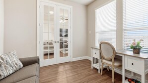 Una habitación con piso de madera, un escritorio blanco con silla, un sofá y una puerta con paneles de vidrio.