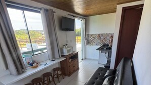 A modern living room with a black sofa, wooden ceiling, a TV mounted on the wall, a kitchen area with a microwave, and a view of the outdoors through large windows. - Pousada Mahara (Imbituba)