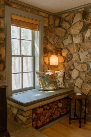 A stone wall, a window with blinds, a bench with cushions, a wall-mounted lamp, and a small wooden table.