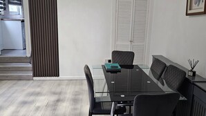 Un espace de salle à manger moderne avec une table en verre, des chaises noires, un lustre et une vue sur une cuisine et un escalier.