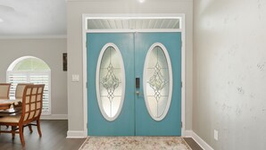 A room with a teal double door, a dining area with wooden chairs, and a patterned rug. - Hugh Bear Haven: Pool, Tiki Bar, Kayaks, Dog-Friendly (Crystal River)