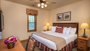 Ein Hotelzimmer mit Bett, zwei Nachttischlampen, Deckenventilator, Fenster mit Jalousien, Holz-Kopfteil und einem kleinen Tisch mit Blumenvase.