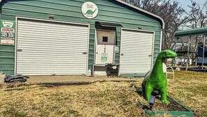 Un bâtiment vert avec deux portes de garage, une enseigne pour la station-service Sinclair Dino, et une statue de dinosaure verte devant.