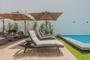 Uma área à beira da piscina com espreguiçadeiras, guarda-sóis e uma churrasqueira.