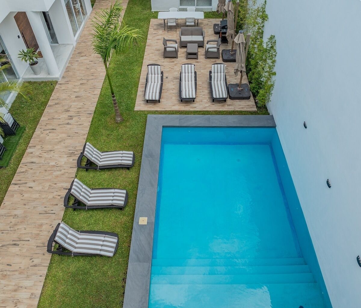 Una piscina en la azotea con sillones, una mesa y sillas, rodeada por una terraza de madera y un área con césped.