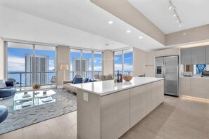 A modern kitchen with a large island, a dining area with a glass coffee table, and a view of the cityscape through large windows. - Skyline Penthouse  Private Balcony & Ocean Views (Hallandale Beach)