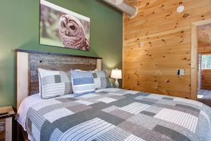 A bedroom with a wooden headboard, a bed with a patterned bedspread, pillows, a wall-mounted picture of an owl, and a wooden wall panel.