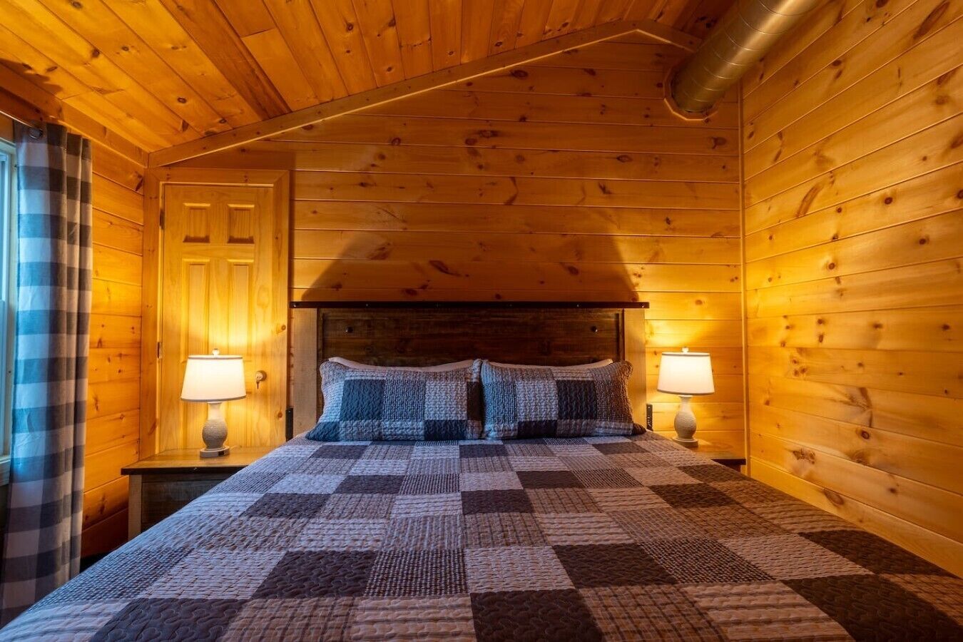 A cozy wooden cabin room with a checkered bedspread, two bedside lamps, and a window with plaid curtains.
