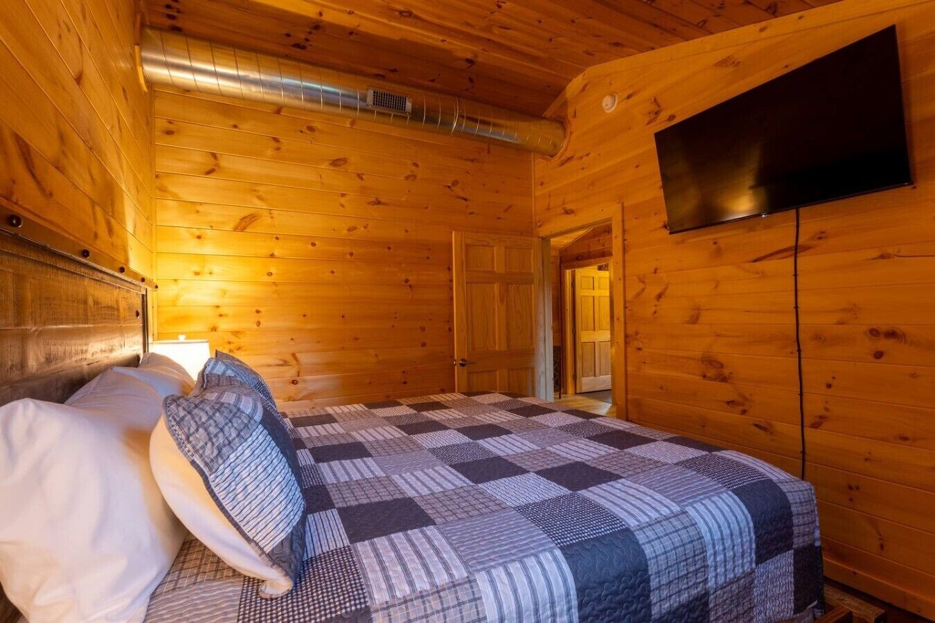 A wooden cabin room with a bed, a television mounted on the wall, and a visible door leading to another room.