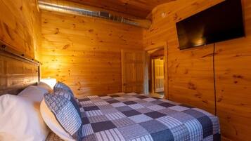 A wooden cabin room with a bed, a television mounted on the wall, and a visible door leading to another room.