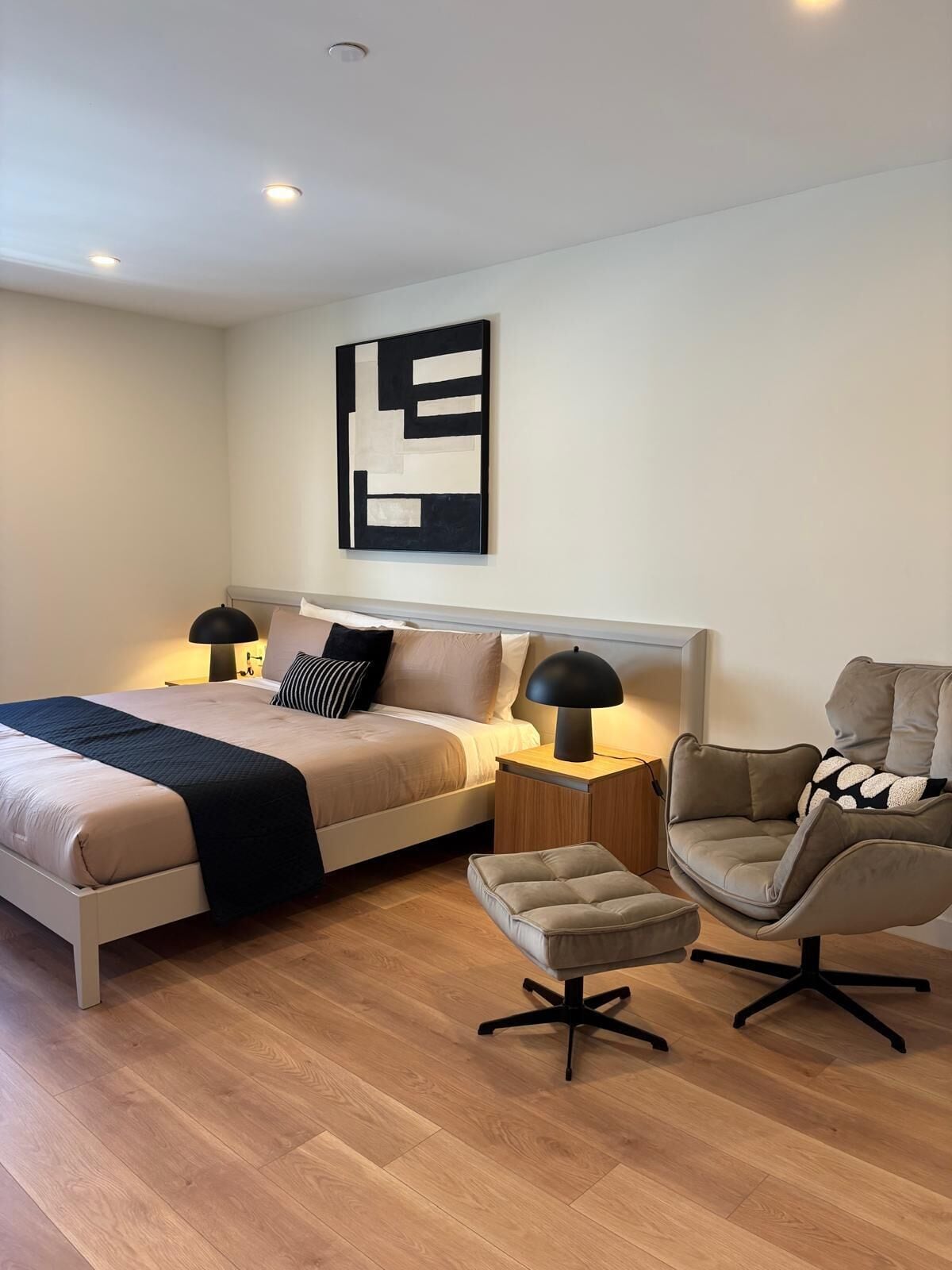 Un dormitorio moderno con cama, lámparas de noche, una silla y una obra de arte en la pared.