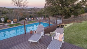 A wooden deck with a pool, lounge chairs, and a fenced area with chairs and tables.